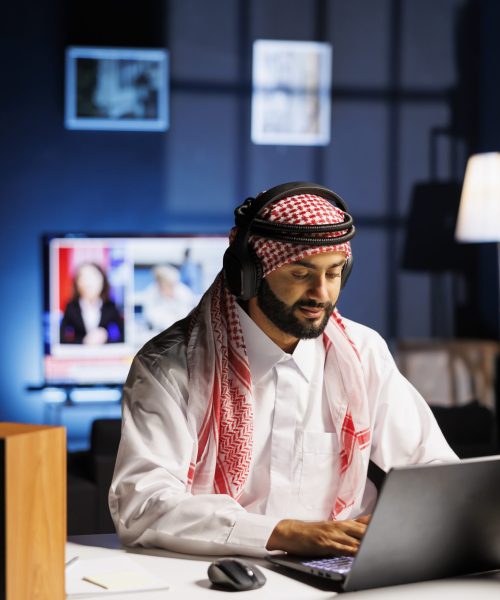 Arab peMiddle Eastern man in traditional attire working indoors, using a laptop for browsing while engaged in video call, using wireless headphones. Image shows male Arab person utilizing modern technology.rson using modern technology arbab of Trivanet TechnolMiddle Eastern man in traditional attire working indoors, using a laptop for browsing while engaged in video call, using wireless headphones. Image shows male Arab person utilizing modern technology.ogies